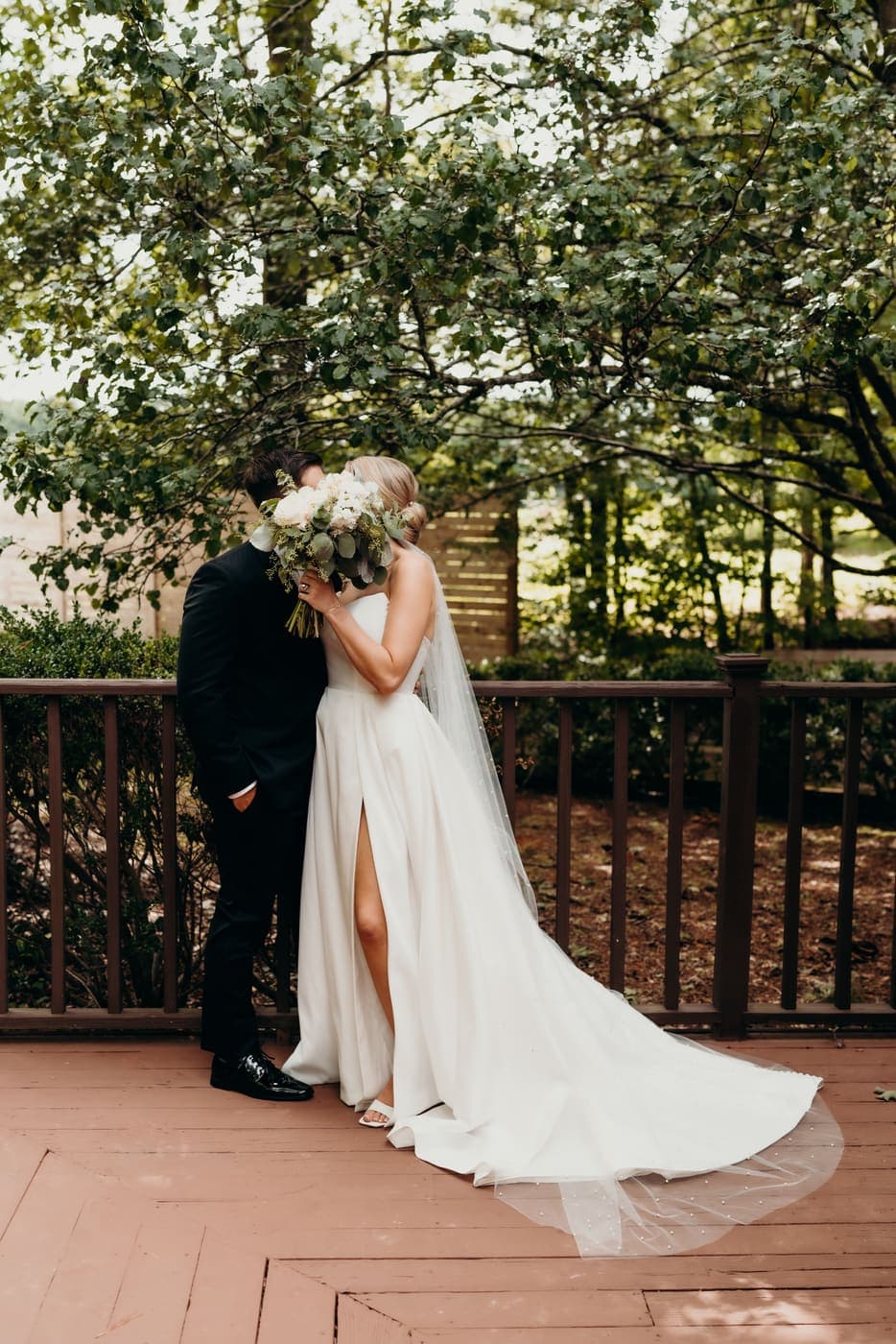 A couple close together, holding a bouquet beneath the trees.
